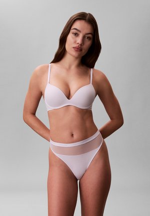Donna che indossa un reggiseno color lavanda chiaro e slip a vita alta abbinati, in piedi con le mani dietro la schiena su uno sfondo grigio uniforme.