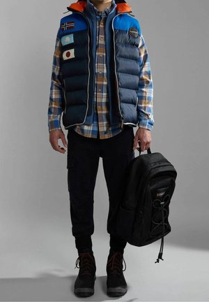 Persona che indossa un gilet imbottito blu e arancione sopra una camicia a quadri, pantaloni cargo neri e stivali, mentre tiene uno zaino nero per il manico.