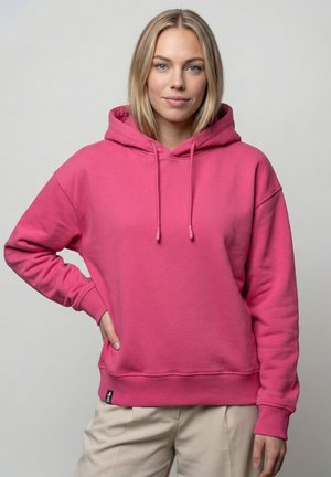 Junge Frau mit schulterlangem blondem Haar trägt eine knallig pinke Kapuzenjacke und hellbeige Hose, steht mit einer Hand in der Hüfte.