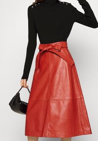 Czerwona skórzana spódnica o długości do połowy łydki z wiązanym paskiem w talii, gładką fakturą i prostymi liniami. Dopasowana do czarnego, prążkowanego golfu i torebki.