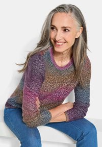 Femme aux longs cheveux gris portant un pull en tricot multicolore et un jean bleu, assise les bras croisés, souriante et regardant à droite.