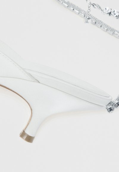 Sapato branco de salto alto com bico pontiagudo, costura visível e base pequena com salto quadrado, ao lado de fios de joias prateadas com strass.