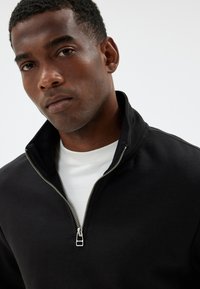 Black zip-up sweatshirt with a high collar, made of soft fabric, featuring a silver zipper and layered white t-shirt underneath.
