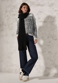 Pull en tricot avec un motif géométrique noir et blanc, associé à un jean large noir et à une épaisse écharpe noire. Baskets blanches avec accents noirs.
