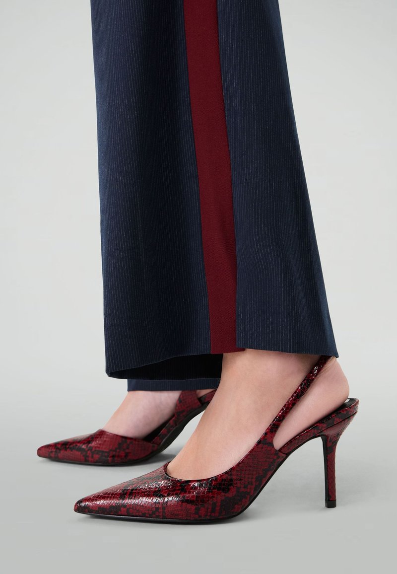 Chaussures à talons aiguilles à imprimé serpent rouge avec un bout pointu et une finition brillante, associées à un pantalon bleu marine avec une rayure bourgogne.