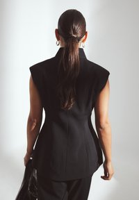 Schwarzer taillierter ärmelloser Blazer mit strukturierten Schultern, taillierter Taille und glatter Textur; getragen mit passenden schmal geschnittenen Hosen.