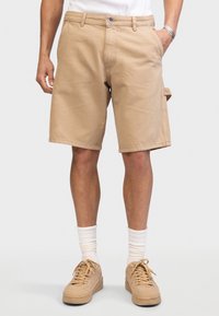 Mann trägt beiges knielanges Shorts, weiße Socken und beige Turnschuhe, steht mit einer Hand in der Tasche vor einem einfarbigen Hintergrund.