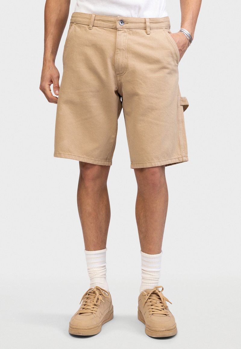 Mann trägt beiges knielanges Shorts, weiße Socken und beige Turnschuhe, steht mit einer Hand in der Tasche vor einem einfarbigen Hintergrund.