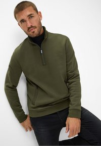 Felpa zip-up verde scuro con maniche lunghe, colletto rialzato e tasca laterale. Tessuto liscio, design aderente, abbinata a jeans neri.
