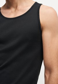 Nahaufnahme einer Person, die ein schwarzes ärmelloses Tanktop trägt, das die obere Brust, Schulter und den Arm vor neutralem Hintergrund zeigt.
