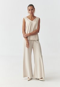 Beige ilmanhiha blouse, jossa on V-pääntie, yhdistettynä leveälahkeisiin housuihin. Sujuva, kevyt kangas; kärjen muotoiset beige korot; pitkät roikkuvat korvakorut.