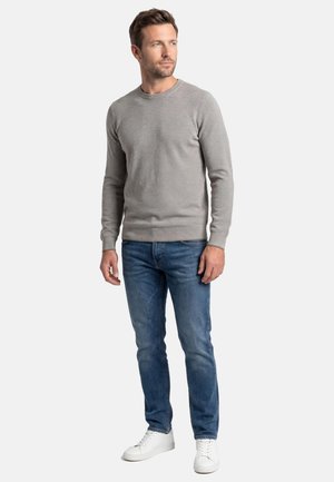 Man staat en draagt een grijze trui met lange mouwen, blauwe jeans en witte sneakers, kijkt opzij tegen een effen achtergrond.