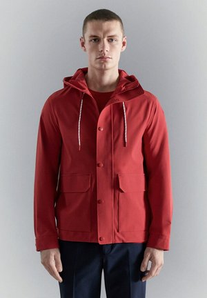 Joven con una chaqueta roja con capucha y bolsillos delanteros, y pantalones negros, posando frente a un fondo liso de color gris claro.