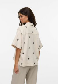 Mujer con cabello largo y oscuro, vestida con una camisa suelta de mangas cortas color beige con patrones de diamantes negros y pantalones gris claro, de perfil.