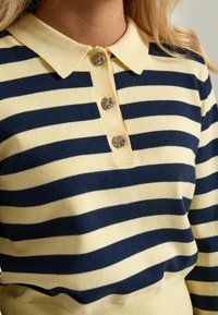Námořní modré a žluté pruhované pletené polo tričko s límečkem, se třemi kovovými knoflíky s květinovým vzorem, měkkou texturou a přiléhavým střihem.