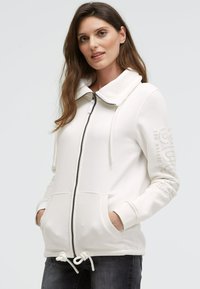 Weiße Zip-Hoodie mit hohem Kragen, Kordelzugbund, Fronttaschen und Text-Detail am Ärmel. Hergestellt aus weichem, strukturiertem Stoff.