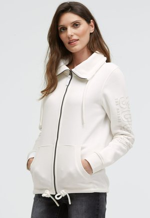 White zip-up hoodie with a high collar, drawstring waist, front pockets, and text detail on the sleeve. Made from soft, textured fabric.