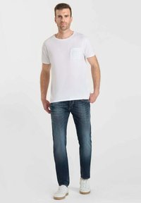 Camiseta blanca de manga corta con un bolsillo texturizado en el pecho, combinada con pantalones vaqueros azul oscuro y zapatillas blancas, de pie contra un fondo neutral.