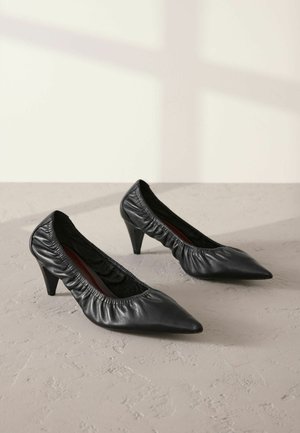 Escarpins en cuir noir avec un bout pointu, texture plissée autour de la cheville et un talon fin. Fini lisse sur la surface supérieure.