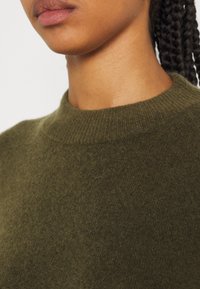 Maglione a maglia verde oliva con un colletto alto e a costine, che mostra una texture morbida e una vestibilità aderente. Design minimalista e senza motivi visibili.
