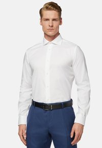 Chemise blanche à manches longues avec un col pointu, boutonnée sur le devant et d'une texture lisse, associée à un pantalon bleu marine et une ceinture noire.
