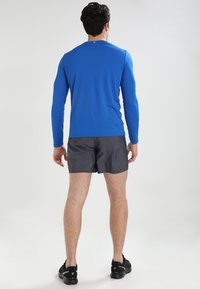 Camisa atlética de manga larga azul, hecha de tela liviana, combinada con pantalones cortos grises. Unos zapatos deportivos negros completan el atuendo.