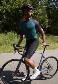 Cycliste portant un maillot ajusté teal et violet et un short noir avec un logo. Vélo moderne avec des roues noires et des freins à disque. Cadre estival.
