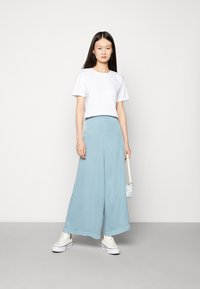 Camiseta branca cropped combinada com calças largas de tom azul claro. A modelo usa ténis brancos e segura uma pequena bolsa com padrão.