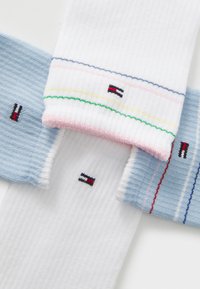 Tre par med ribbede bomullssokker i hvit og blå, med stripete detaljer og en liten logo-patch i rød, svart og hvit.