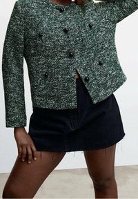 Veste vert texturée à double boutonnage avec des boutons noirs et un col arrondi, associée à une jupe en denim noir. Manches courtes et longueur raccourcie.