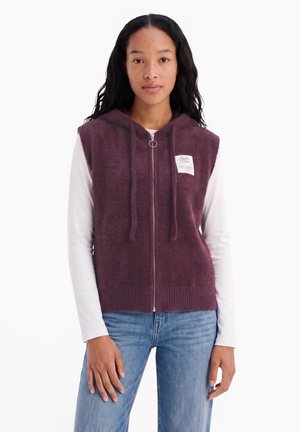 Giovane donna che indossa un gilet con cappuccio senza maniche color melograno peloso sopra una camicia bianca a maniche lunghe e jeans blu, in piedi davanti a uno sfondo bianco.