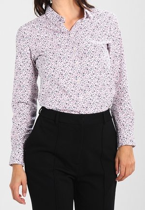 Femme portant un chemisier fleuri à manches longues, de couleur claire, rentré dans un pantalon noir taille haute et ajusté.