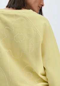 Hellgelbes Sweatshirt mit weißen gestickten Blumenmustern auf dem Rücken, aus weichem Textil mit einem entspannten Design.