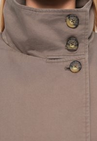 Manteau beige à col montant avec trois grands boutons ronds fixés sur le côté et une petite fente de poche horizontale.