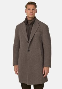 Cappotto a spina di pesce marrone con colletto a punta, chiusura a un bottone e due tasche laterali. Tessuto testurizzato e sovrapposto con una giacca interna scura.