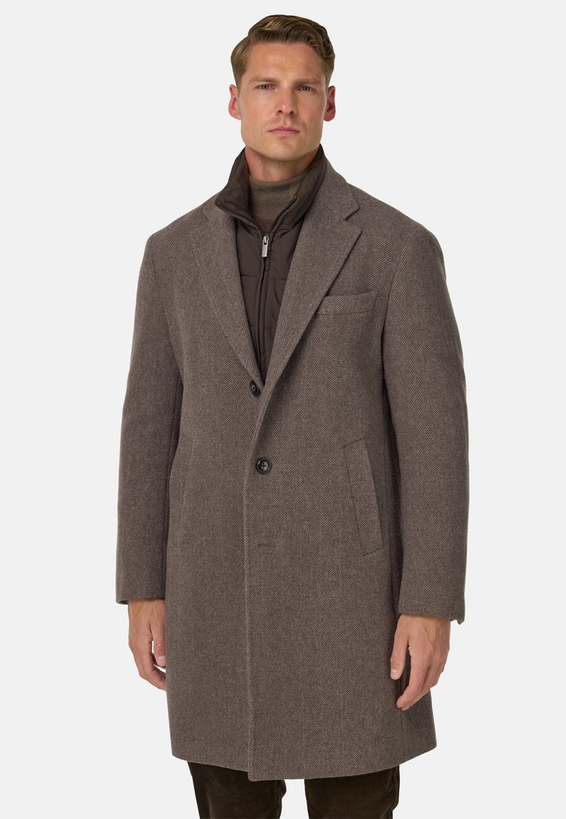 Cappotto a spina di pesce marrone con colletto a punta, chiusura a un bottone e due tasche laterali. Tessuto testurizzato e sovrapposto con una giacca interna scura.