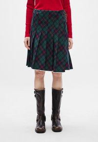 Πράσινη καρό φούστα με κόκκινες λεπτομέρειες, πλισέ σχέδιο και φ fringe τελείωμα. Συνδυάζεται με καφέ δερμάτινες μπότες μέχρι το γόνατο με αγκράφες.