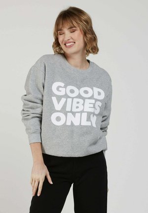 Jeune femme souriante, les yeux fermés, portant un sweat-shirt gris avec l'inscription "GOOD VIBES ONLY" et un pantalon noir, sur un fond uni.