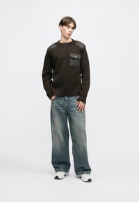 Barna pulóver válldíszekkel, mellzsebes kivitelben. Laza kék farmernadrággal és fehér sportcipővel viselve, egyszínű háttér előtt.