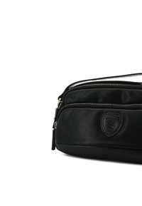 Bolsa de cuero negro con dos compartimentos con cremallera, acabado texturizado, parche con logo y acentos en hardware plateado. Forma rectangular compacta.
