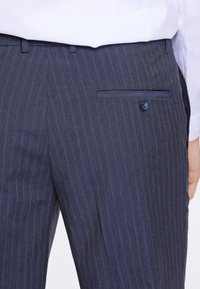 Mörkblå randiga byxor i ett strukturerat tyg, med en bakficka prydd med en blå knapp och bälteshällor.