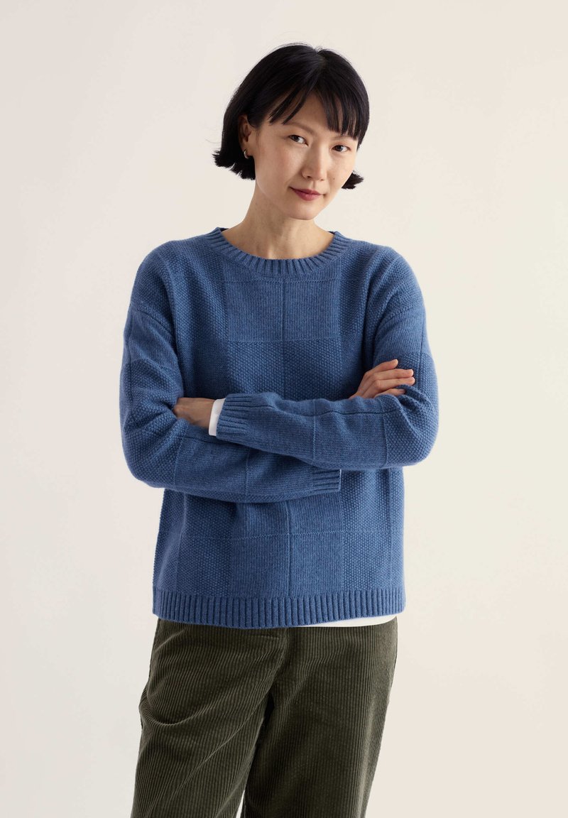 Femme aux cheveux noirs courts portant un pull bleu et un pantalon en velours côtelé vert, debout les bras croisés devant un fond clair uni.