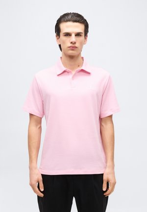 Νέος άνδρας στέκεται με το πρόσωπο προς τα εμπρός, φορώντας ένα ανοιχτό ροζ κοντομάνικο πόλο πουκάμισο και μαύρο παντελόνι, μπροστά σε ένα απλό φόντο.