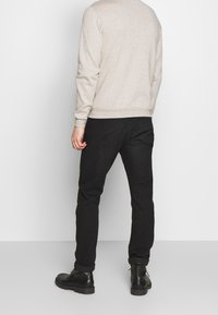 Hombre vistiendo un suéter beige de manga larga, jeans slim negros y botas negras de cordones, de pie de espaldas contra un fondo blanco.