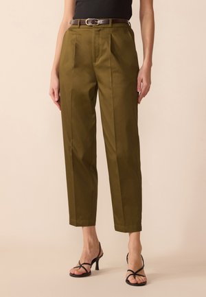 Femme portant un pantalon plissé taille haute vert olive avec une ceinture marron et des sandales à talons noires à brides, debout devant un fond neutre.