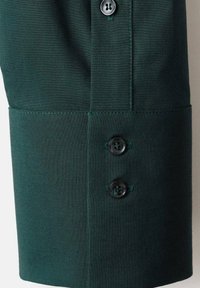 Dark green fabric with a smooth texture, featuring three black buttons and contrasting green stitching at the cuff section.