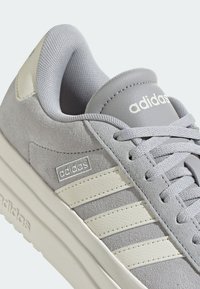 Grå mockaskor med vita läderstreck, vadderad tunga och texturerad snörning. Har logotyp på sidan.