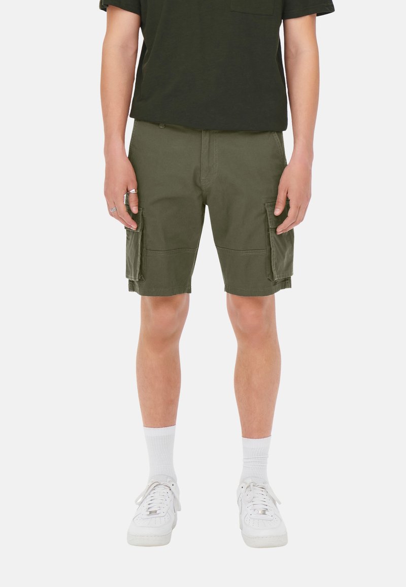 Pantalones cortos cargo verde oliva con dos grandes bolsillos laterales y un ajuste relajado, combinados con una camiseta negra y zapatillas blancas.