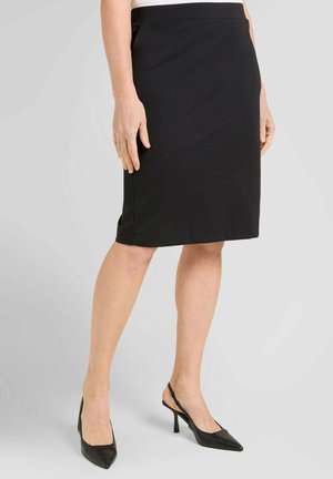 Donna che indossa una gonna a tubino nera al ginocchio abbinata a tacchi slingback neri con punta appuntita, in piedi davanti a uno sfondo grigio semplice.