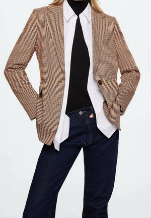 Person trägt braunen karierten Blazer, weißes Hemd mit Knöpfen, schwarzen Rollkragenpullover und dunkelblaue Jeans, Hände in den Taschen, vor schlichtem Hintergrund.